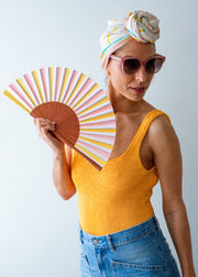 Woman holding a colorful paper fan, wearing sunglasses and a yellow top with a white headscarf.