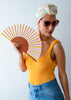 Woman holding a colorful paper fan, wearing sunglasses and a yellow top with a white headscarf.
