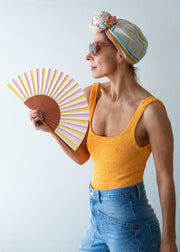 Woman wearing a colorful headscarf, yellow tank top, and blue jeans, holding a striped fan against a light background.