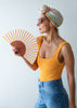 Woman wearing a colorful headscarf, yellow tank top, and blue jeans, holding a striped fan against a light background.