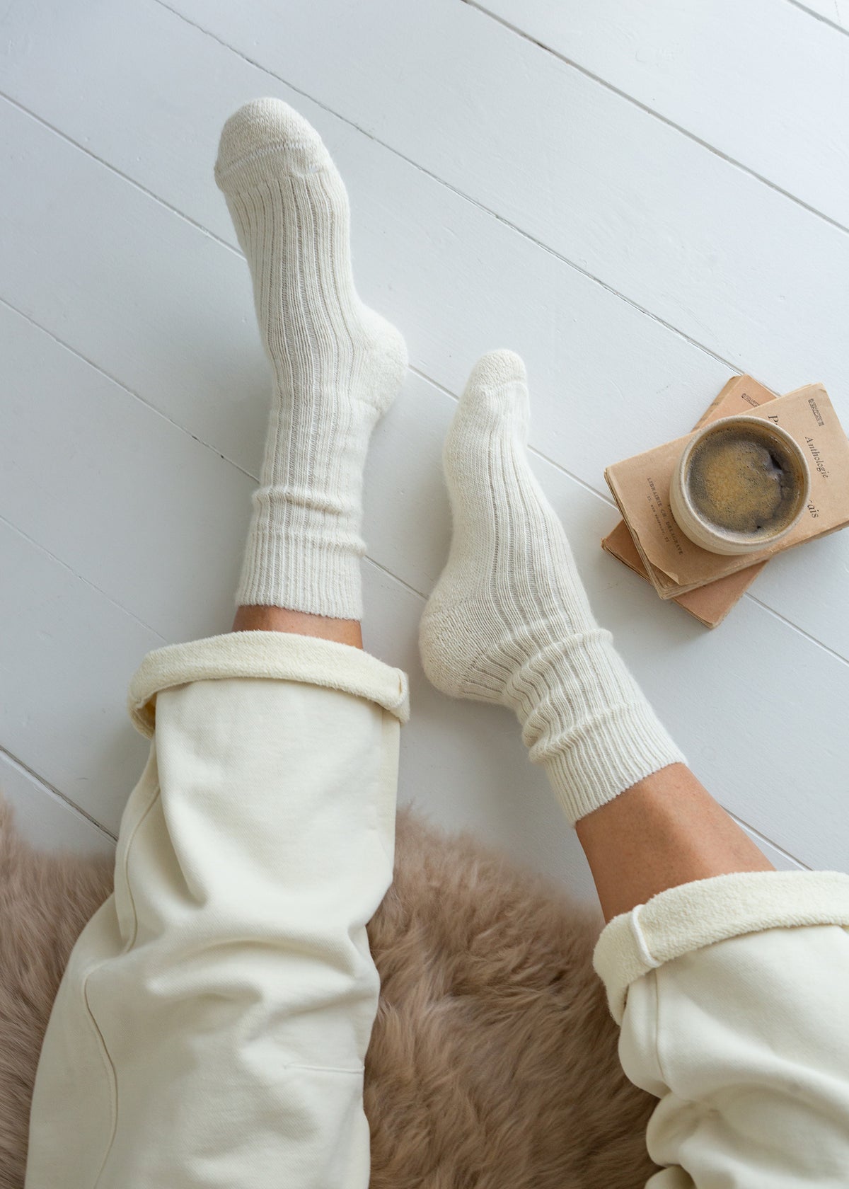 A top-down view of a person wearing cream-colored ribbed crew alpaca socks and matching sweatpants, sitting on a brown faux fur rug with a cup of coffee on wooden coasters nearby, set against a white wooden floor.