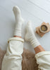 A top-down view of a person wearing cream-colored ribbed crew alpaca socks and matching sweatpants, sitting on a brown faux fur rug with a cup of coffee on wooden coasters nearby, set against a white wooden floor.