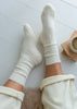 A top-down view of a person's legs wearing off-white ribbed knit crew alpaca socks and matching cream pants, resting on a white wooden floor next to a small cup and notebook.