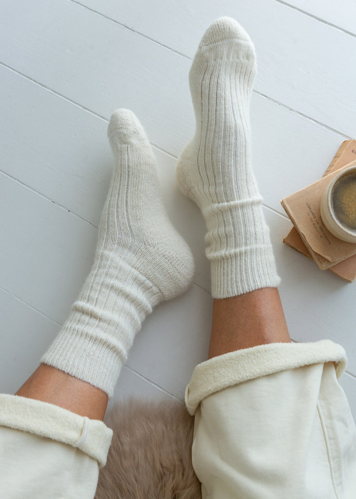 A top-down view of a person's legs wearing off-white ribbed knit crew alpaca socks and matching cream pants, resting on a white wooden floor next to a small cup and notebook.