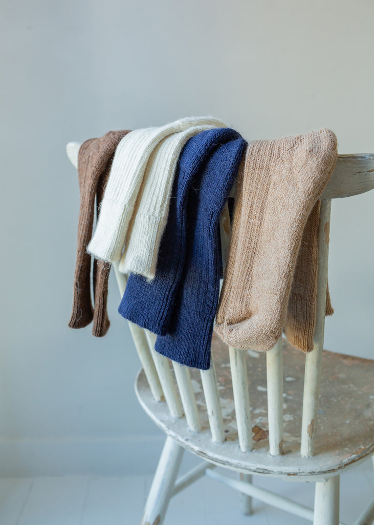 Four ribbed knit alpaca socks in brown, cream, navy, and beige draped over the back of a distressed white wooden chair against a soft light gray wall.