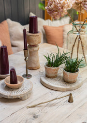 A styled tabletop arrangement with dark burgundy taper candles in wooden holders and a candle snuffer , small potted green plants on round stone trays, a brass candle holder, and dried pink flowers in a glass vase on a light wood table.