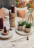 A styled tabletop arrangement with dark burgundy taper candles in wooden holders and a candle snuffer , small potted green plants on round stone trays, a brass candle holder, and dried pink flowers in a glass vase on a light wood table.