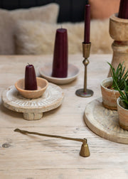 A styled tabletop scene with dark burgundy taper candles in wooden holders, a brass candle snuffer , a brass scythe candle, and small potted green plants on a light wood surface.