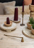 A styled tabletop scene with dark burgundy taper candles in wooden holders, a brass candle snuffer , a brass scythe candle, and small potted green plants on a light wood surface.