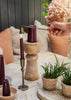 A hand holds a brass candle snuffer over a dark purple taper candle in a rustic wooden holder, set on a light wood table with other candles, potted green plants, and neutral-toned cushions in the background.