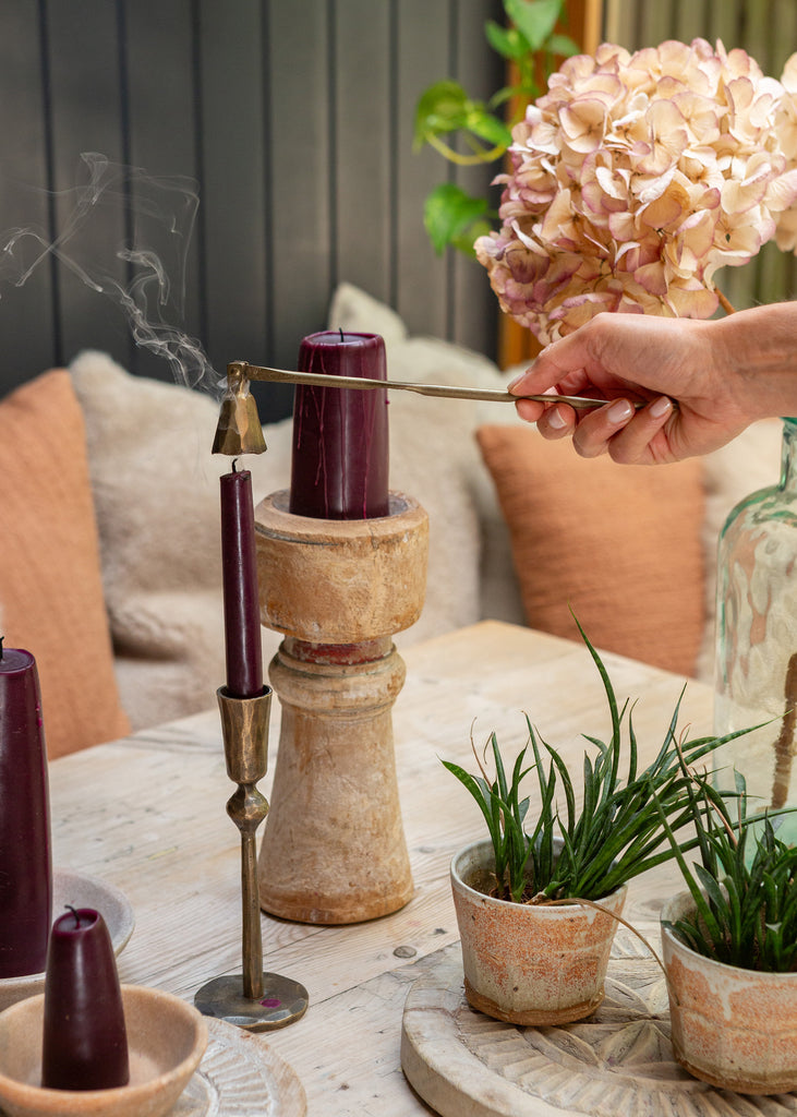 A hand holds a long metal candle snuffer over a dark purple taper candle in a rustic wooden holder, set on a light wood table with other candles, potted green plants, and a bouquet of pale pink hydrangeas in the background.