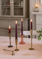 Four lit taper candles in brass and pink glass candlesticks arranged on a table in a softly lit room with a candle snuffer.