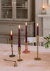 Four lit taper candles in brass and pink glass candlesticks arranged on a table in a softly lit room with a candle snuffer.