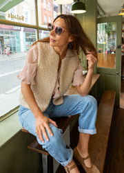 A woman wearing a beige sherpa vest over a light pink blouse, blue jeans, and brown shoes sits on a wooden bench in a cafe setting.