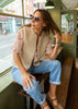 A woman wearing a beige sherpa vest over a light pink blouse, blue jeans, and brown shoes sits on a wooden bench in a cafe setting.