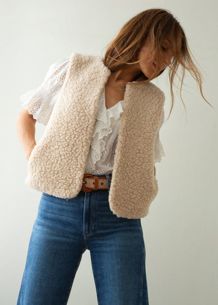 A woman wearing a cream-colored, textured sherpa fleece vest over a white lace blouse and blue jeans, photographed against a plain light gray background.