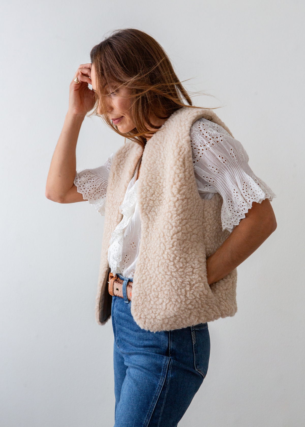 A woman wearing a beige sherpa fleece vest over a white eyelet blouse and blue jeans, standing against a plain light gray background.
