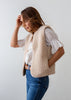A woman wearing a beige sherpa fleece vest over a white eyelet blouse and blue jeans, standing against a plain light gray background.