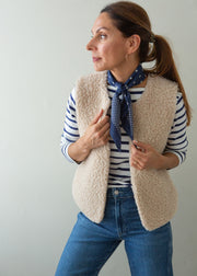 A woman wearing a beige sherpa fleece vest over a navy and white striped long-sleeve shirt, accessorized with a navy patterned neck scarf and blue jeans, posing against a light gray wall.