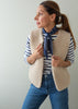 A woman wearing a beige sherpa fleece vest over a navy and white striped long-sleeve shirt, accessorized with a navy patterned neck scarf and blue jeans, posing against a light gray wall.