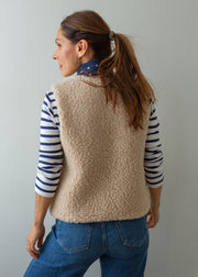 A woman shown from the back wearing a beige sherpa fleece vest over a navy and white striped long-sleeve shirt, paired with blue jeans, against a plain light gray background.