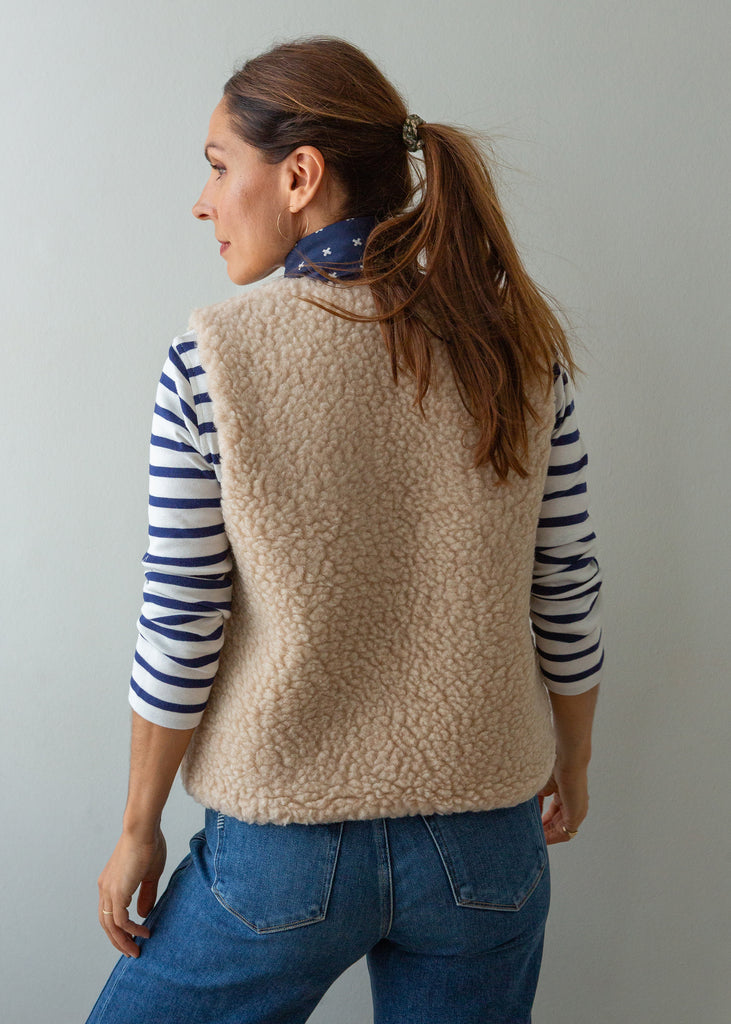 A woman shown from the back wearing a beige sherpa fleece vest over a navy and white striped long-sleeve shirt, paired with blue jeans, against a plain light gray background.