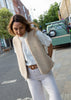 A woman wearing a cream-colored teddy fleece vest over a white eyelet blouse and white trousers, standing on a city street.