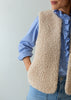 A person wearing a cream-colored sherpa fleece vest over a blue and white striped shirt and blue jeans, shown from the chest down against a plain light gray background.