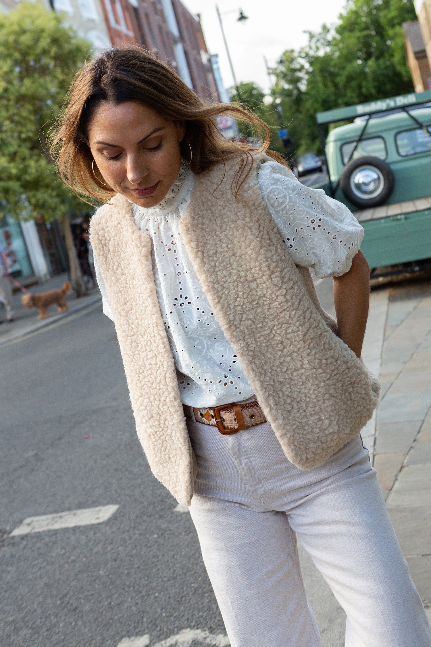 A woman wearing a cream-colored textured tmerino gilet over a white eyelet blouse, paired with white trousers and a Guatemalan belt, standing on a city street.