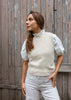 A woman wearing a cream-colored ribbed knit sweater tank over a white eyelet puff-sleeve blouse and light gray trousers, standing in front of a weathered wooden door.