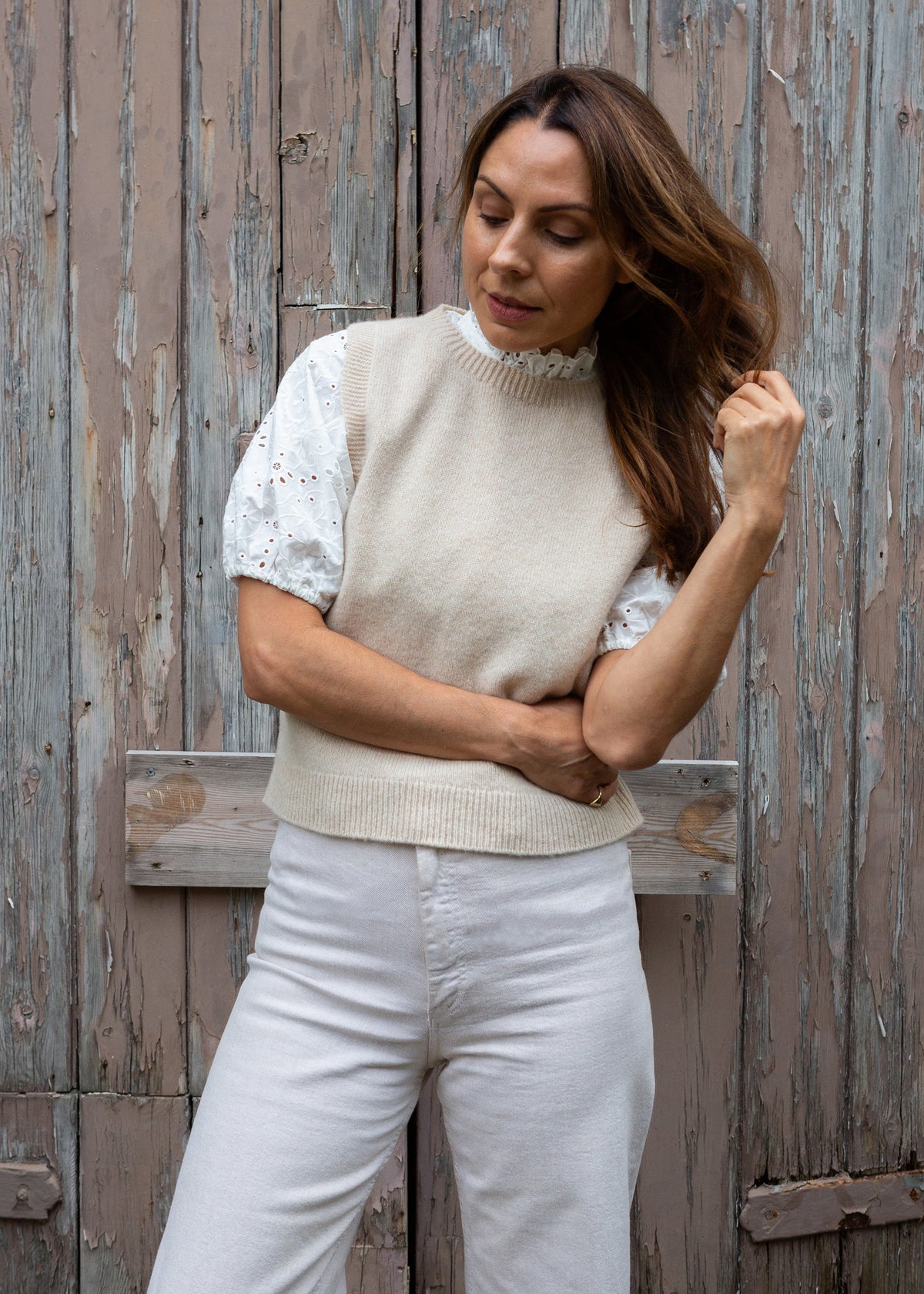 A woman wearing a cream knit sweater vest over a white lace-sleeve top and white trousers, standing against a weathered wooden door.