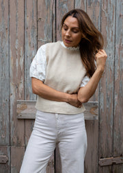 A woman wearing a cream knit sweater vest over a white lace-sleeve top and white trousers, standing against a weathered wooden door.