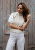 A woman wearing a cream knit sweater vest over a white lace-sleeve top and white trousers, standing against a weathered wooden door.