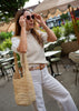 A woman in a white lace-sleeve top, cream knit sweater vest, and white trousers carries a large woven straw tote bag while posing outdoors at a cafe with patio umbrellas.