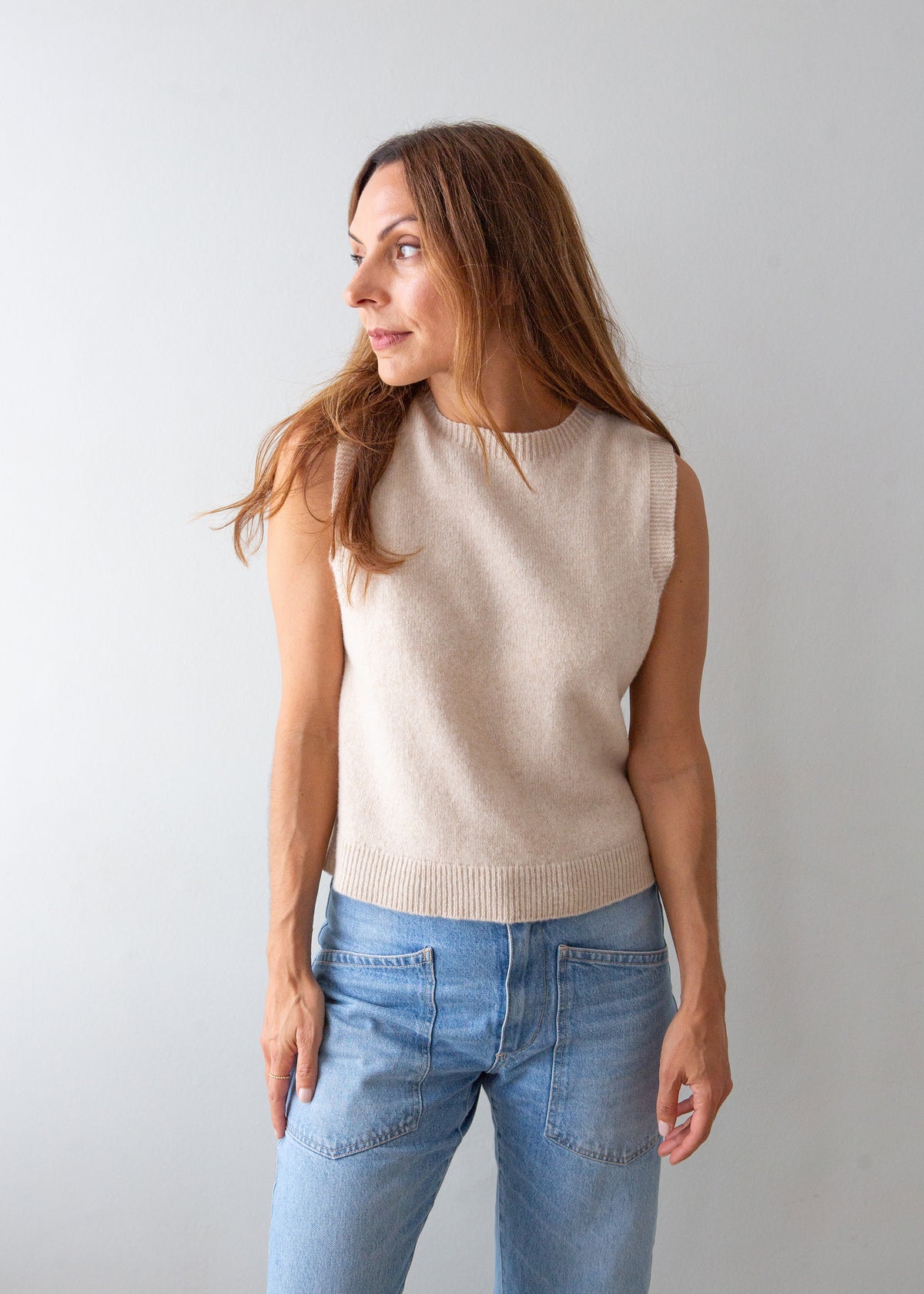 A woman wearing a sleeveless beige knit sweater vest and light blue jeans, standing against a plain light gray background.