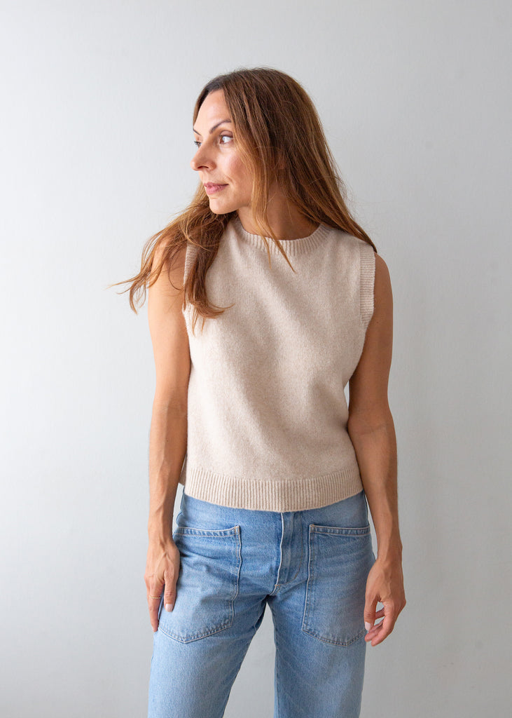 A woman wearing a sleeveless beige knit sweater vest and light blue jeans, standing against a plain light gray background.