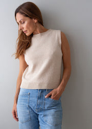 A woman wearing a sleeveless cream knit sweater vest and light blue jeans, standing against a plain gray wall.