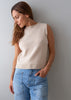 A woman wearing a sleeveless cream knit sweater vest and light blue jeans, standing against a plain gray wall.