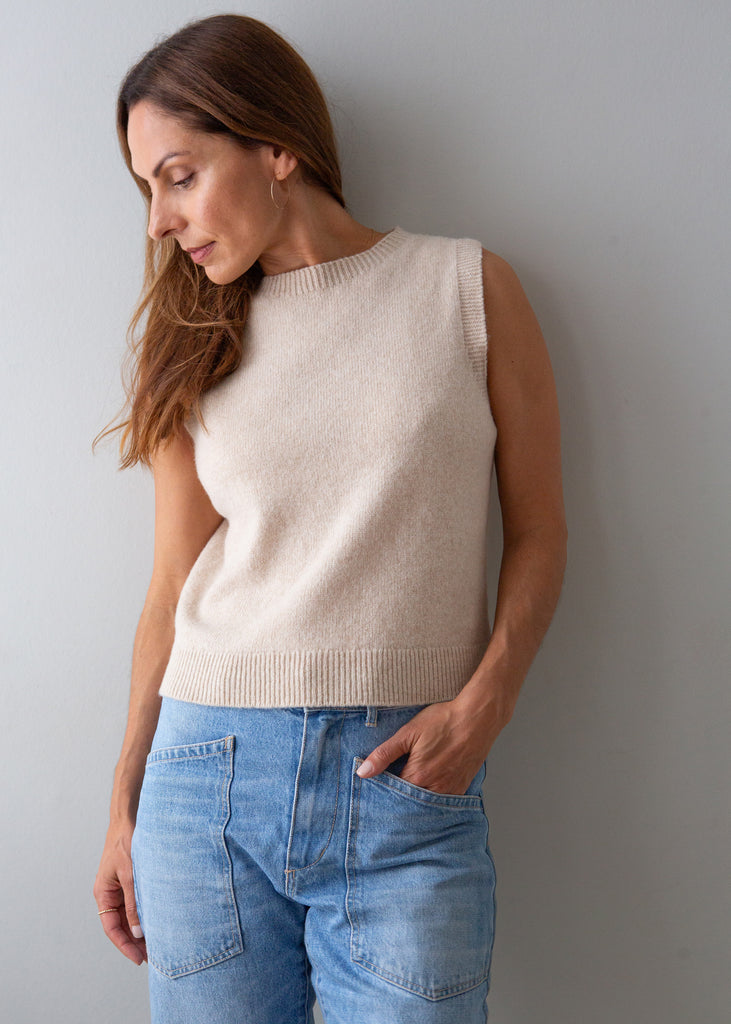 A woman wearing a sleeveless cream knit sweater vest and light blue jeans, standing against a plain gray wall.