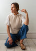 A woman kneels on a light wood floor against a white wall, wearing a beige knit sweater vest with white lace flutter sleeves, blue jeans, and brown Mary Jane shoes.
