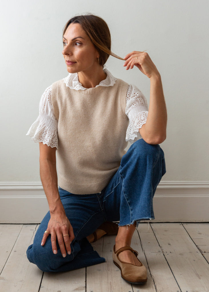 A woman kneels on a light wood floor against a white wall, wearing a beige knit sweater vest with white lace flutter sleeves, blue jeans, and brown Mary Jane shoes.