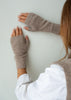 A person wearing taupe knit fingerless gloves, shown from the side against a plain light gray wall.