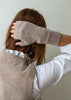 A person shown from the back wearing a taupe knit sweater vest over a white long-sleeve top, with one hand raised to adjust a matching taupe knit fingerless glove. The background is a plain light gray wall.