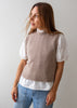 A woman wearing a taupe knit sweater vest over a white eyelet blouse with puffed short sleeves, paired with light blue jeans, photographed against a plain light gray background.