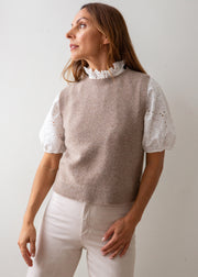 A woman wearing a taupe knit sweater vest over a white eyelet puff-sleeve top with a ruffled collar, paired with light beige pants, photographed against a plain light gray background.