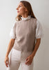 A woman wearing a taupe knit sweater vest over a white eyelet puff-sleeve top with a ruffled collar, paired with light beige pants, photographed against a plain light gray background.