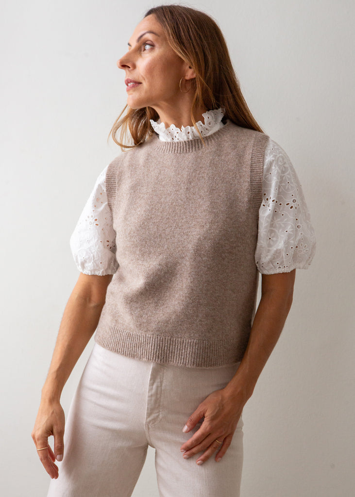 A woman wearing a taupe knit sweater vest over a white eyelet puff-sleeve top with a ruffled collar, paired with light beige pants, photographed against a plain light gray background.