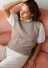 A woman wearing a beige knit sweater vest over a white eyelet blouse and cream trousers, sitting on a dusty pink couch.