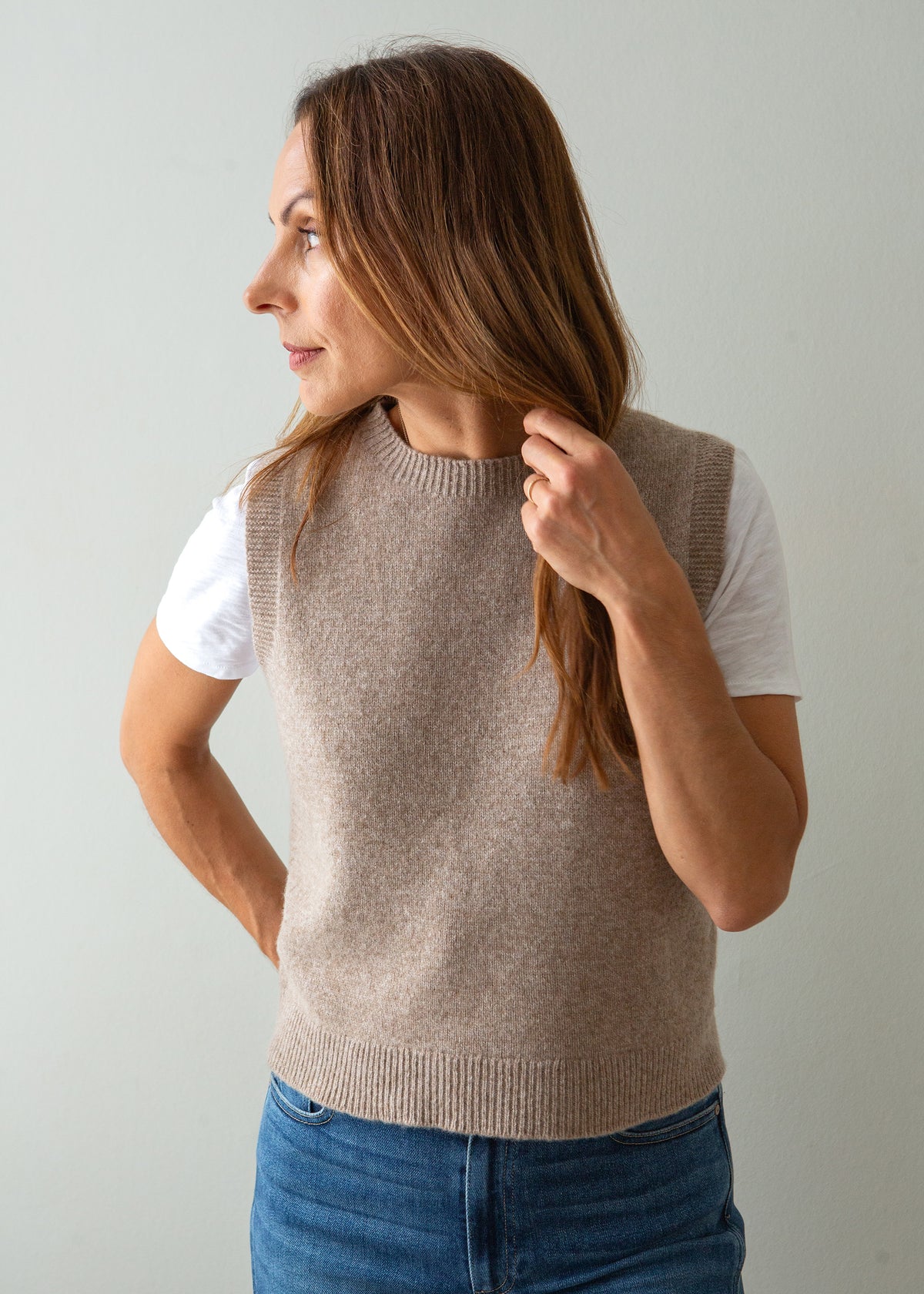 A woman wearing a sleeveless knit sweater vest in a light taupe-beige color over a white t-shirt and blue jeans, shown from a side profile against a plain light gray background.