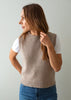 A woman wearing a sleeveless knit sweater vest in a light taupe-beige color over a white t-shirt and blue jeans, shown from a side profile against a plain light gray background.